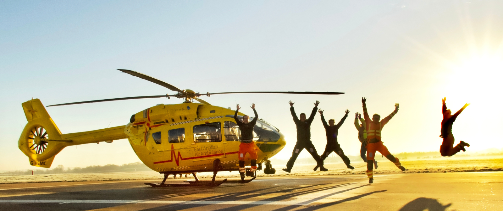 Crew jumping next to yellow, EAAA helicopter at sunset creating shadows on the ground