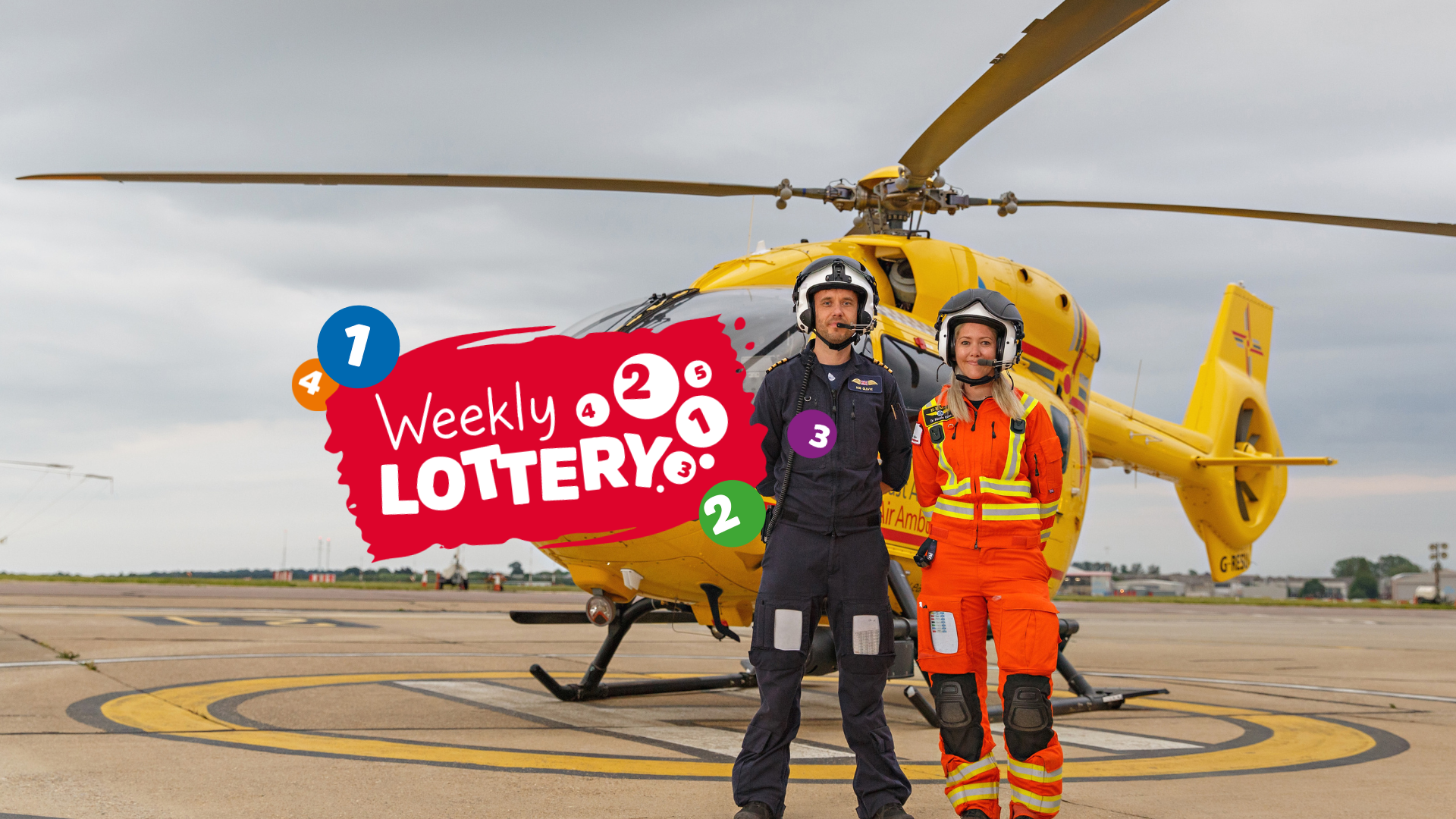 Yellow East Anglian Air Ambulance helicopter with pilot and doctor in flight suits standing in front with red 'weekly lottery' logo